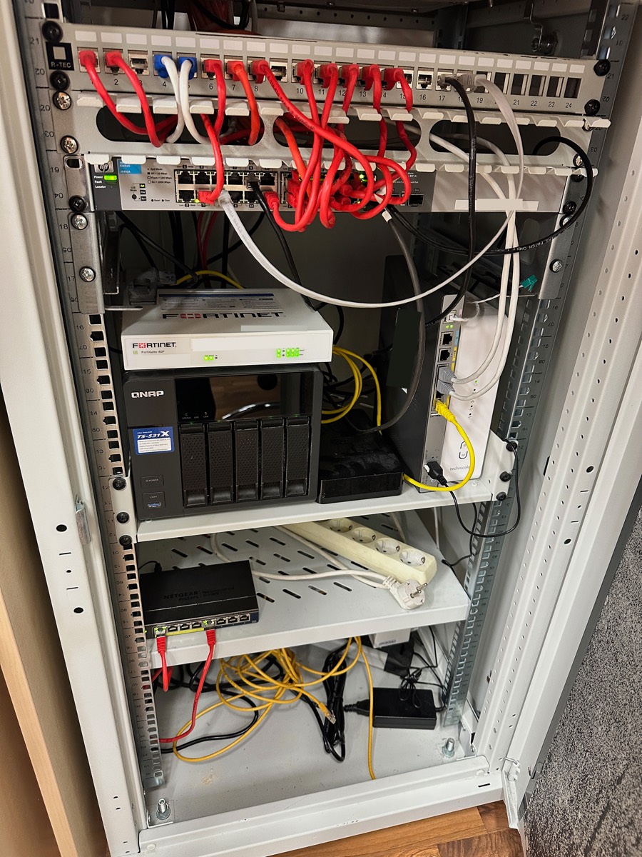 Vorher: der zweite Standort. Auch hier eine FortiGate, dazu QNAP-NAS, drei Switches verschiedener Hersteller und ein Steckdosenverteiler quer über der Ablage.
